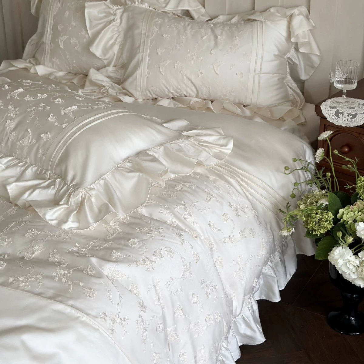 White embroidered bedding set on a bed with a side table and flowers in the background.