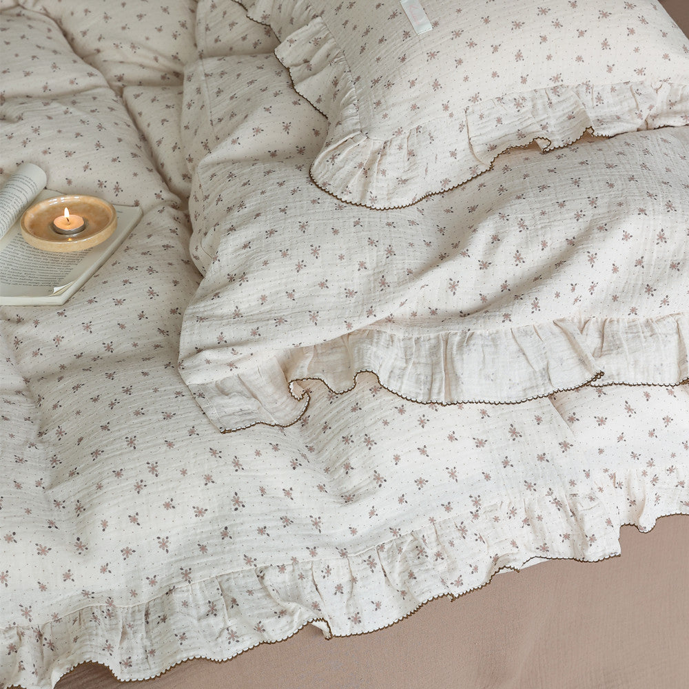 Top-down view of the floral muslin duvet cover and pillow sham, clearly showing the ruffle trim, the light brown floral print, and a small lit candle on the bed.