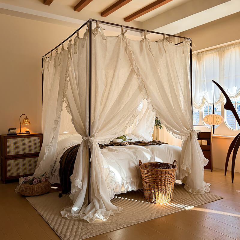 Bedroom with a canopy bed draped in white curtains, wooden floors, and a lamp on a nightstand.