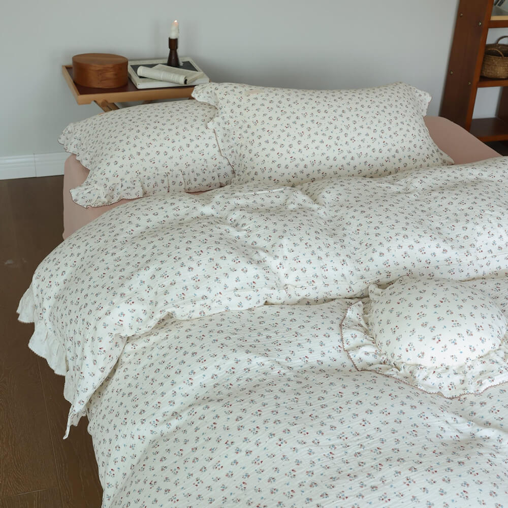Styled shot of the 'Baby's Breath' cotton muslin duvet cover set on a bed, showing the duvet, pillows, and a round decorative pillow, with a candle and book on a bedside table