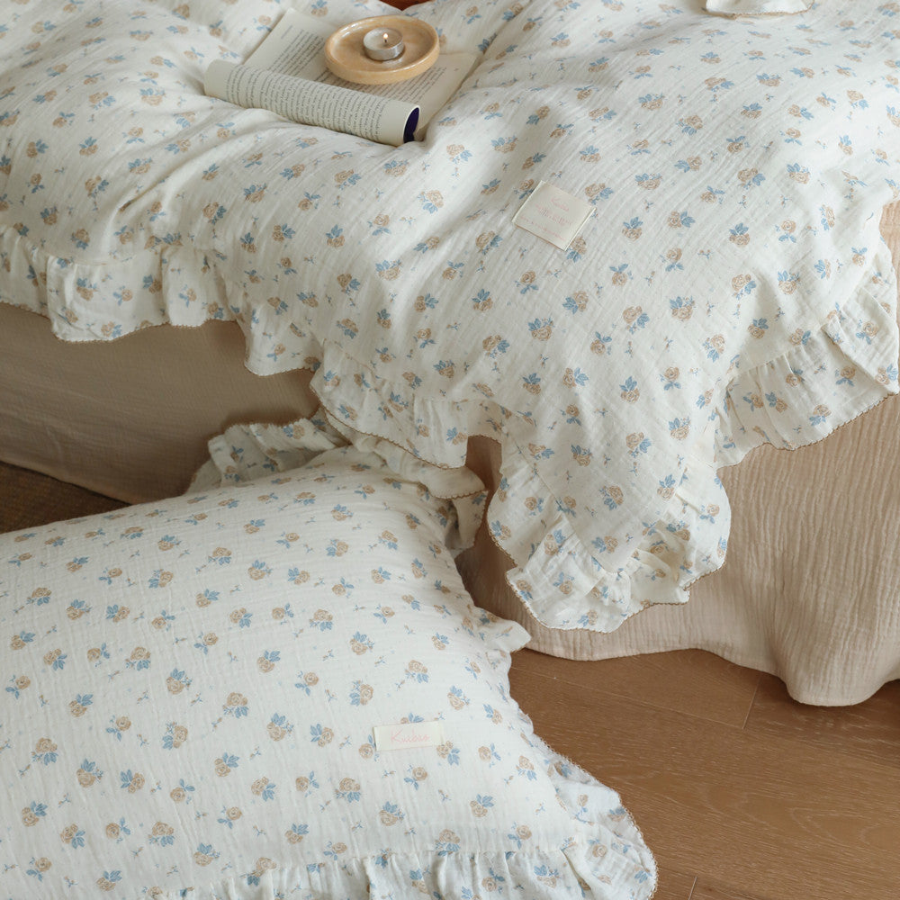  Close-up of a folded vintage rose cotton muslin duvet cover set on a bed. The image highlights the texture of the fabric, the delicate floral print, and the ruffles on the edge of the duvet and a matching pillow, with an open book and lit candle in the foreground.