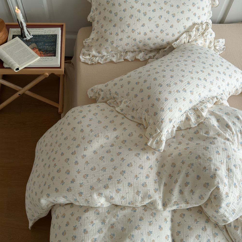 An overhead shot of the bed showing the vintage rose cotton muslin duvet cover and matching pillows with ruffled borders. The light-colored bedding contrasts with the beige bed skirt and wooden side table with a book and candle.