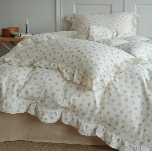 A full view of a bed styled with the vintage rose cotton muslin duvet cover set. The bedding features a light, airy design with floral patterns and ruffled edges on the duvet and pillows. The overall look is cozy and romantic.
