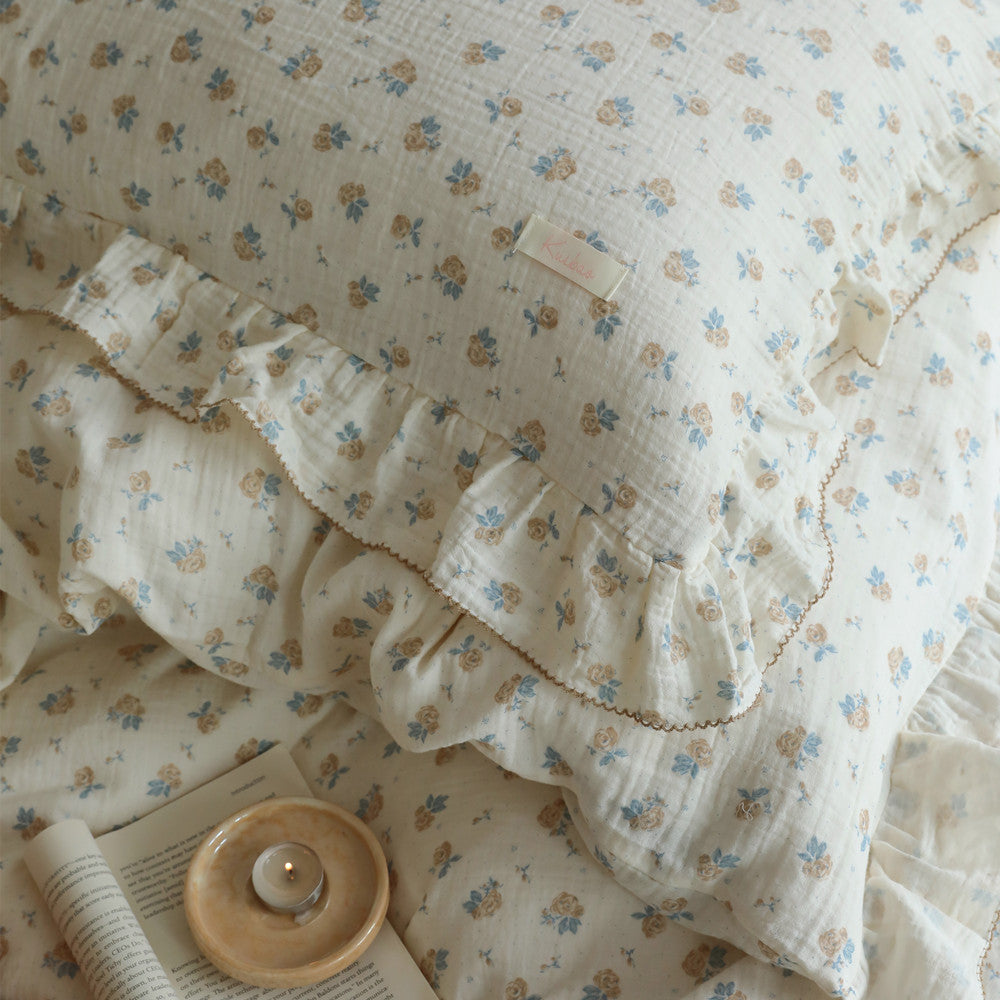 Close-up of a vintage rose patterned cotton muslin duvet cover set with delicate ruffles and a small tag, next to an open book and a lit candle on a small dish. The focus is on the soft texture and floral print of the bedding
