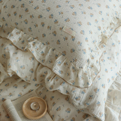 Close-up of a vintage rose patterned cotton muslin duvet cover set with delicate ruffles and a small tag, next to an open book and a lit candle on a small dish. The focus is on the soft texture and floral print of the bedding