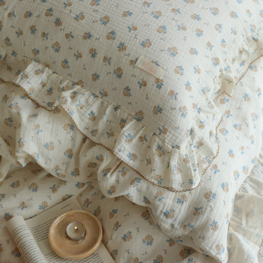 Close-up of a vintage rose patterned cotton muslin duvet cover set with delicate ruffles and a small tag, next to an open book and a lit candle on a small dish. The focus is on the soft texture and floral print of the bedding