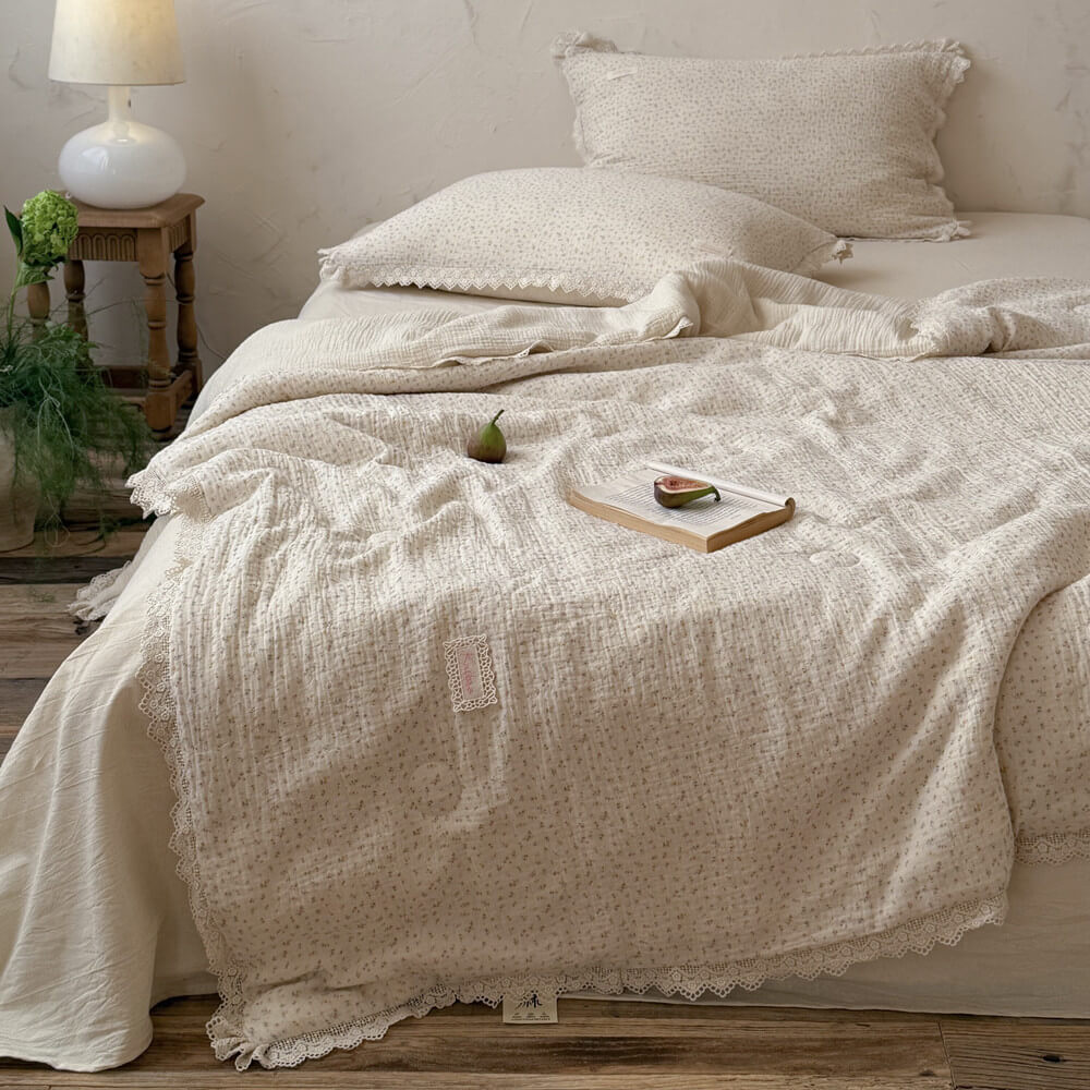 Vintage ditsy floral comforter with delicate lace trim spread on a cozy bed, styled with an open book, fresh figs, and a retro white table lamp for a romantic cottagecore bedroom aesthetic.