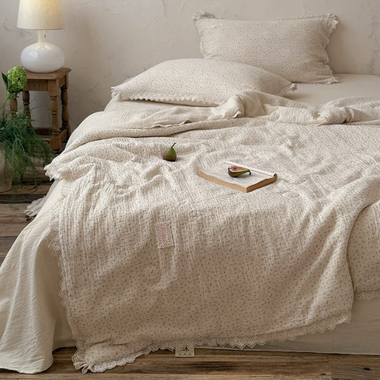 Vintage ditsy floral comforter with delicate lace trim spread on a cozy bed, styled with an open book, fresh figs, and a retro white table lamp for a romantic cottagecore bedroom aesthetic.