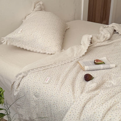 Elegant cream muslin blanket featuring a tiny ditsy flower pattern and vintage lace borders, decorated with a book and fresh fig on a cottage-style bedding display.