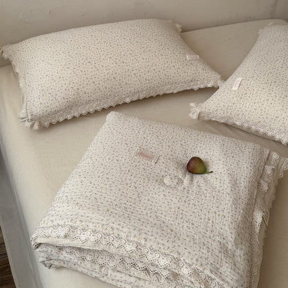 Neat fold of a ditsy floral comforter showing intricate lace trimming, arranged next to matching cotton gauze ruffled pillows and a whole fig fruit in a minimalist rustic bedroom.