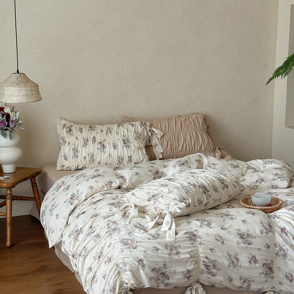 Detailed view of cotton muslin bedding with faded rose floral print, gathered ruching, and multiple ribbon-tie pillow details.