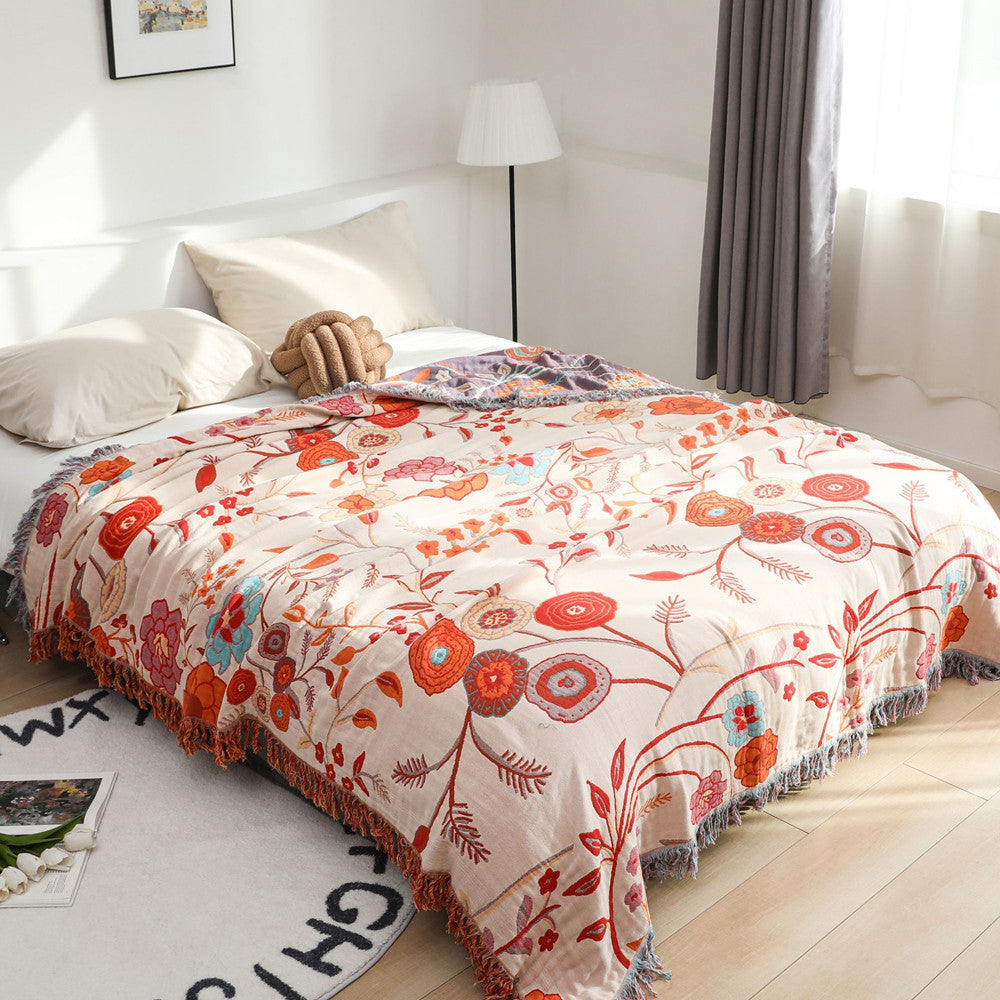 A large floral cotton jacquard throw blanket in a light beige color with vibrant orange, red, and blue flowers draped over a white queen-sized bed. The blanket features decorative fringe around the edges.