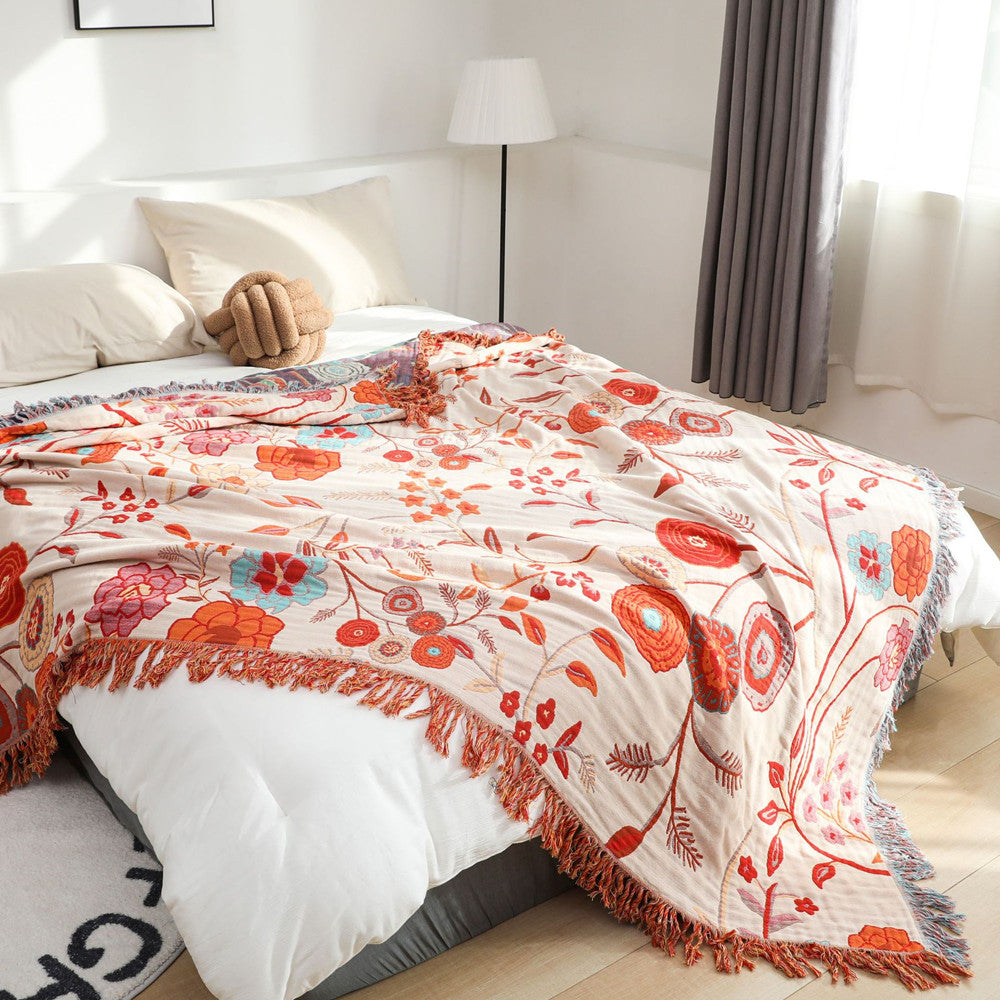 Close-up shot of the reversible floral throw blanket with a tasseled fringe. The jacquard woven cotton fabric in a neutral beige and warm floral pattern is spread across a bed in a bright room.
