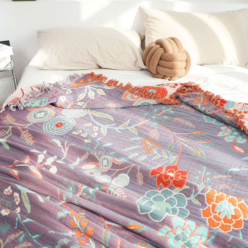 Detailed view of the reverse side of the cotton jacquard throw blanket, showing a dominant deep purple background with the same colorful floral and foliage pattern in orange, teal, and red. The blanket is partially covering a bed.