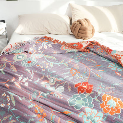 Detailed view of the reverse side of the cotton jacquard throw blanket, showing a dominant deep purple background with the same colorful floral and foliage pattern in orange, teal, and red. The blanket is partially covering a bed.
