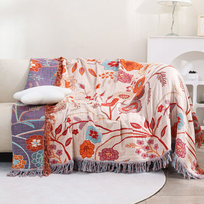 The double-sided floral throw blanket displayed on a light-colored sofa, showcasing both the beige and the deep purple sides. It's a versatile decorative cotton blanket with tassels, suitable for living room decor.