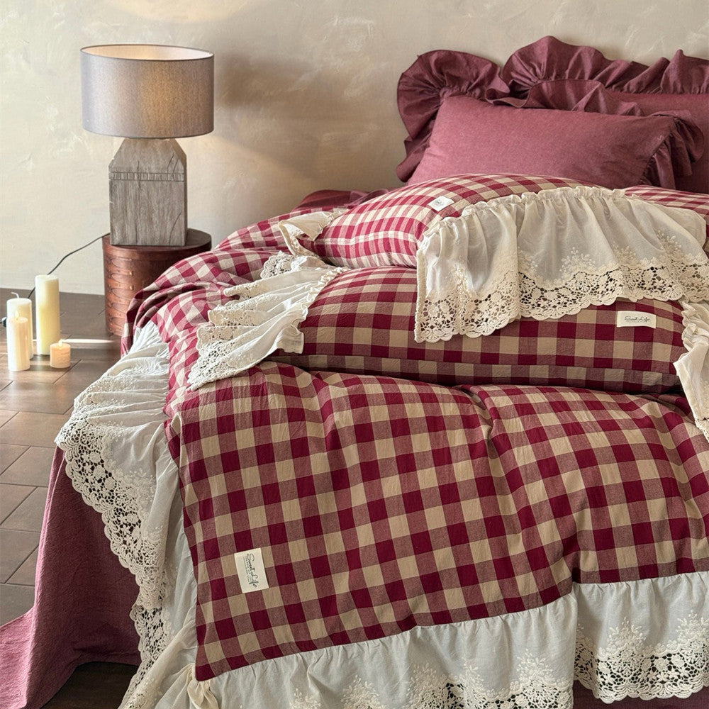 Bed with red and white checkered bedding and lace details, next to a lamp and candles on a wooden floor.