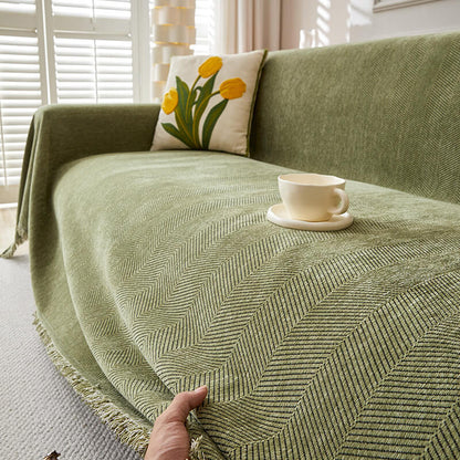 Green herringbone chenille sofa cover with tasseled edges, floral pillow, and cup in bright living room.