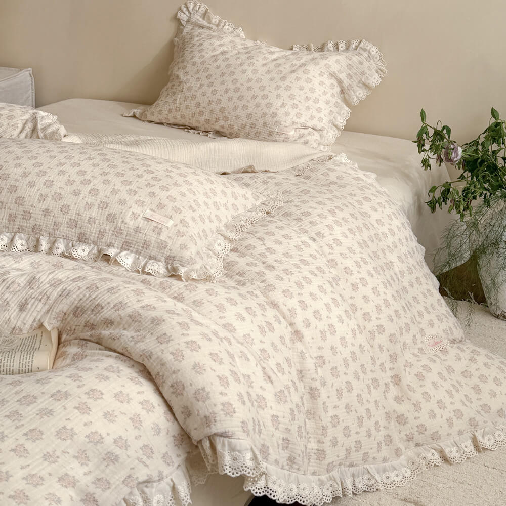 Close-up of a vintage floral duvet cover and matching ruffled pillowcases in muted pink rose patterns with delicate eyelet lace trim detailing.