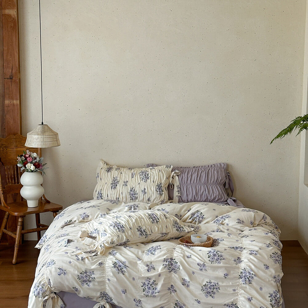 Lavender floral cotton muslin bedding set in a neutral cottage bedroom with ruched pillow shams and ribbon-tie duvet edges.