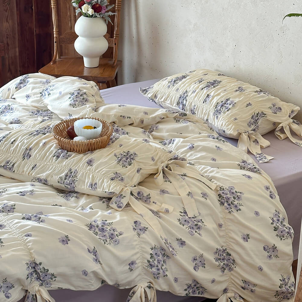 Lavender floral bedding set styled in a warm cottage bedroom with ribbon ties, ruched texture, and wooden decor accents.