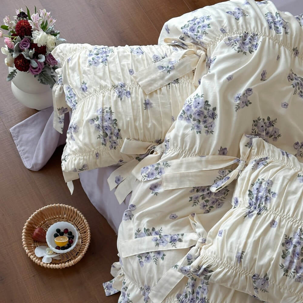 Detailed view of lavender floral cotton muslin bedding with gathered ruching, ribbon ties, and muted purple floral clusters.