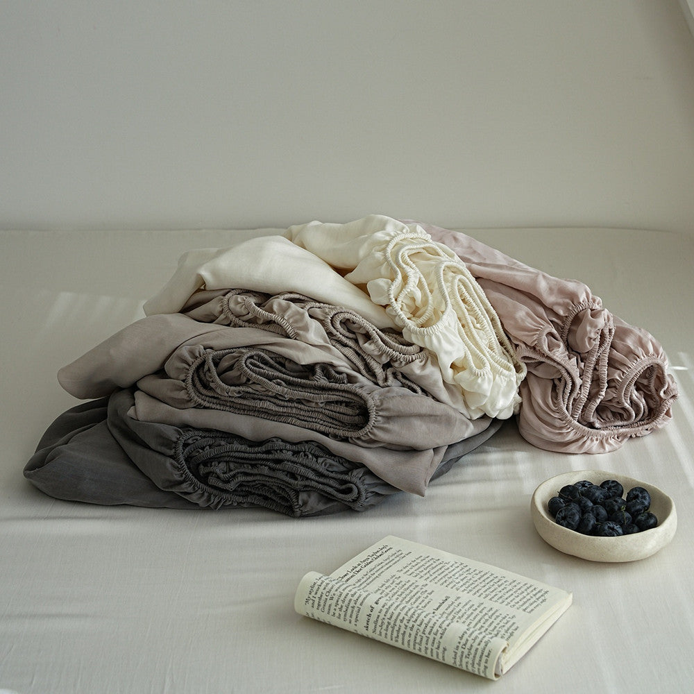 A stack of soft, gathered fabric items in muted neutral tones including gray, taupe, cream, and blush pink, arranged on a bed with a book and a small bowl of blueberries nearby.