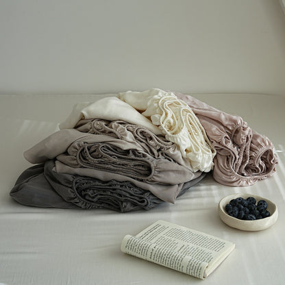 A stack of soft, gathered fabric items in muted neutral tones including gray, taupe, cream, and blush pink, arranged on a bed with a book and a small bowl of blueberries nearby.