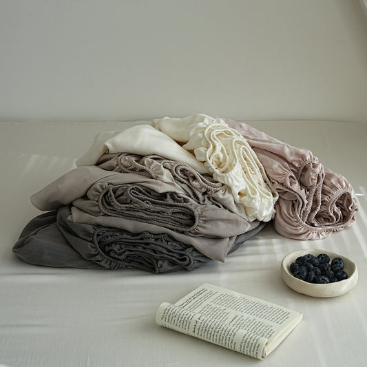A stack of soft, gathered fabric items in muted neutral tones including gray, taupe, cream, and blush pink, arranged on a bed with a book and a small bowl of blueberries nearby.