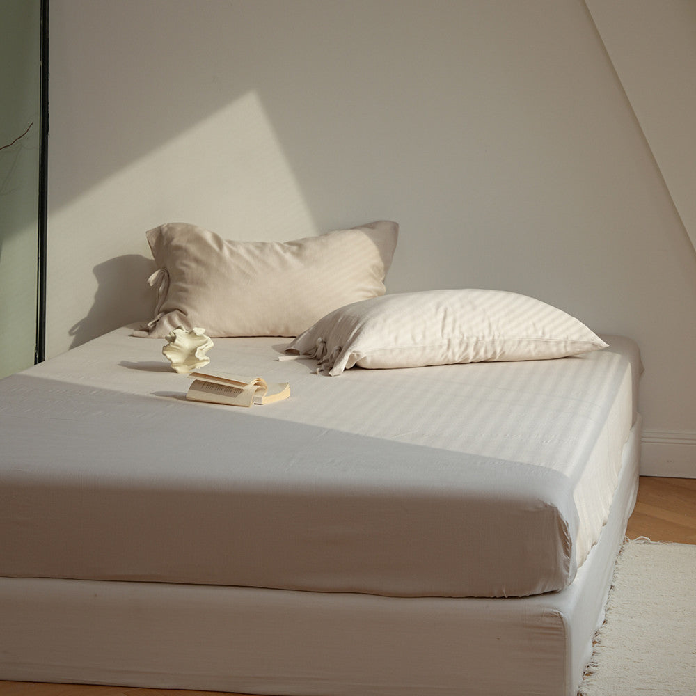 A neatly made bed with a fitted sheet and two pillows in a minimalist beige and cream color palette, set in a room with warm natural light casting diagonal shadows.