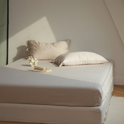 A neatly made bed with a fitted sheet and two pillows in a minimalist beige and cream color palette, set in a room with warm natural light casting diagonal shadows.