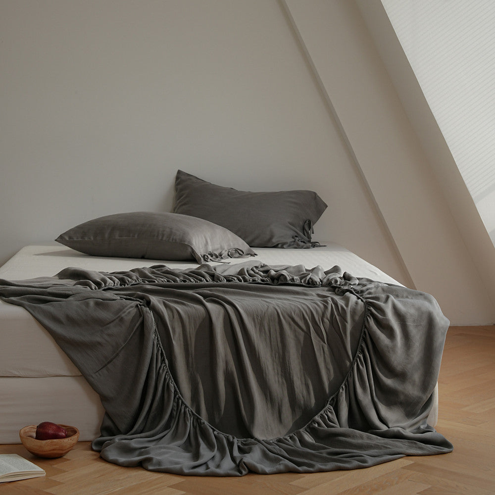 A bed dressed in dark gray bedding, including a draped sheet, pillowcases, and matching pillows, styled in a minimalist attic bedroom with light wood floors and a slanted ceiling.