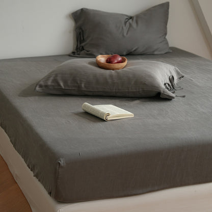 A bed dressed in a dark gray linen sheet and matching pillowcases, styled with a small wooden bowl of fruit and an open book in a softly lit bedroom.