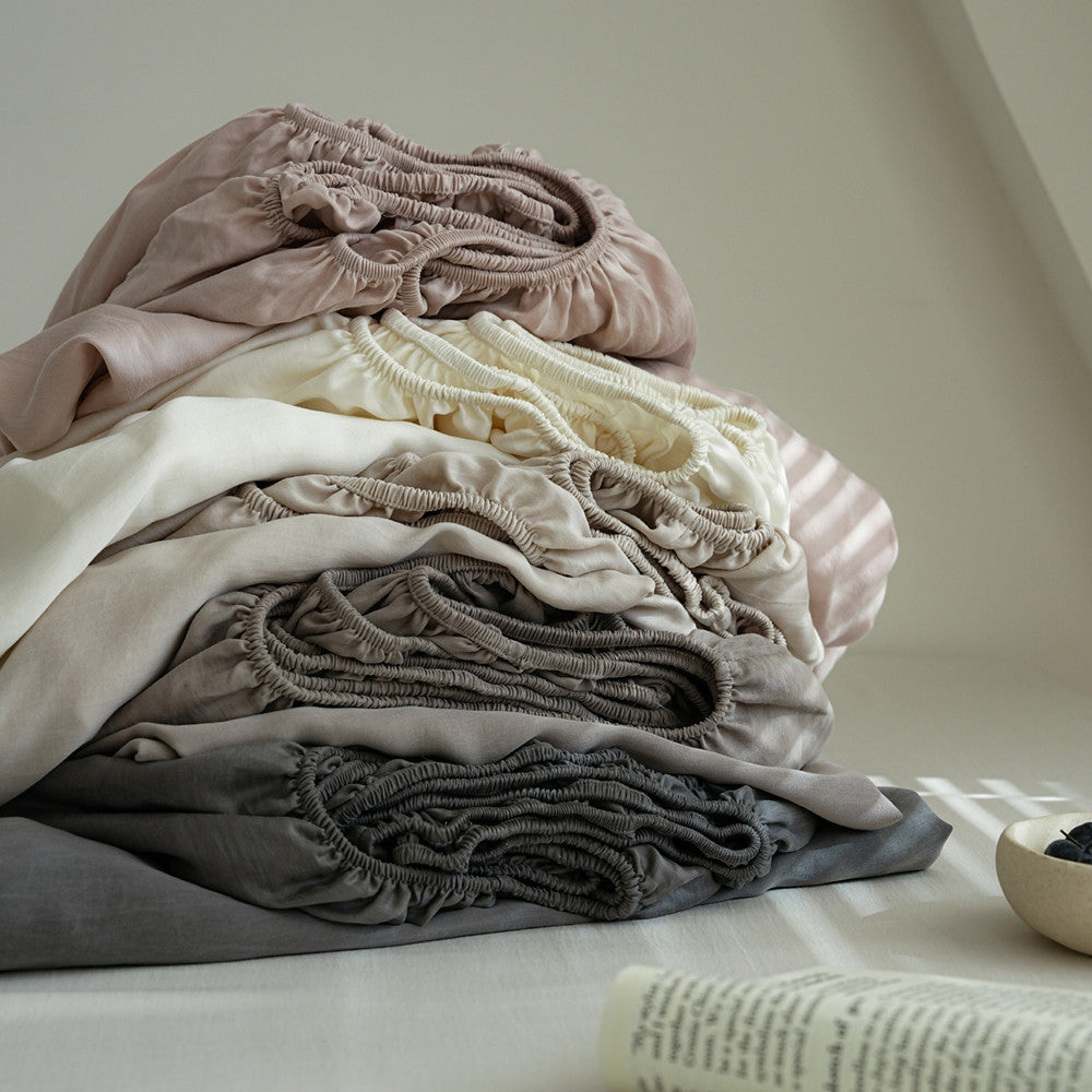 A stack of soft, gathered fabric sheets or bedding in muted neutral tones including dusty pink, cream, taupe, and charcoal gray, styled on a light surface with a book and small bowl in the foreground.