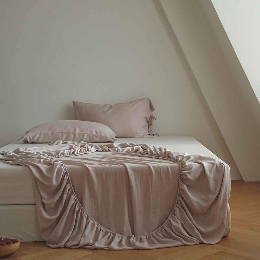 A bed dressed in soft blush pink fitted sheet, including a ruffled slipcover and matching pillows, styled in a minimalist attic bedroom with light wood floors and a slanted ceiling.