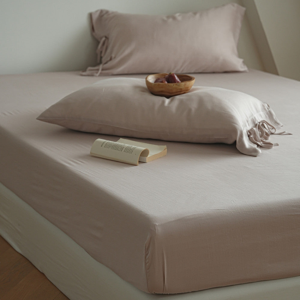 A neatly made bed with a fitted sheet and matching pillowcases in a soft blush-beige tone, styled with two loose pillows, a small wooden bowl, and an open book on the mattress.