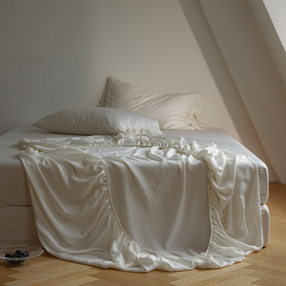 A bed dressed in white bedding, including a white sheet with ruffled edges and two white pillows, set in a softly lit attic bedroom with light wood flooring.