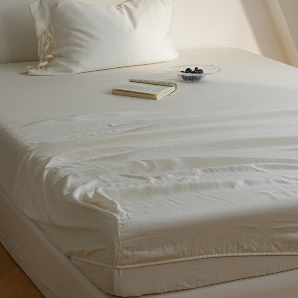 A neatly made bed with white bedding, including a fitted sheet, pillowcases, and a white mattress protector, styled with two pillows, a notebook, and a small glass bowl of blueberries in a softly lit room.
