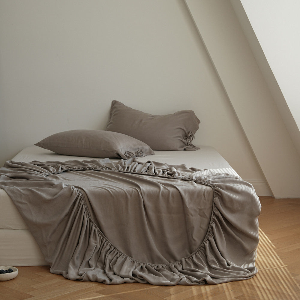 A bed dressed in taupe-gray bedding, including a ruffled slipcover, two matching pillows, and a matching throw blanket, styled in a minimalist attic bedroom with light wood floors.
