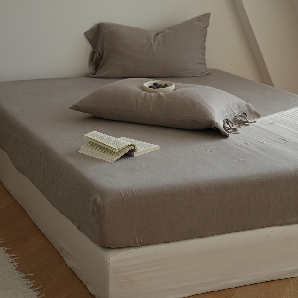 A bed dressed in a taupe-gray fitted sheet and matching pillowcases, styled with two pillows, an open book, and a small dish of blueberries in a bright, minimalist bedroom.
