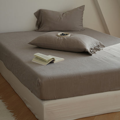 A bed dressed in a taupe-gray fitted sheet and matching pillowcases, styled with two pillows, an open book, and a small dish of blueberries in a bright, minimalist bedroom.