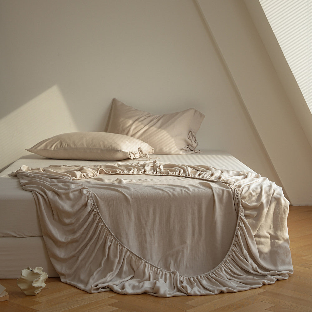 A bed dressed in soft beige bedding, including a ruffled duvet cover and matching pillows, styled in a minimalist attic bedroom with light wood floors and a slanted ceiling.
