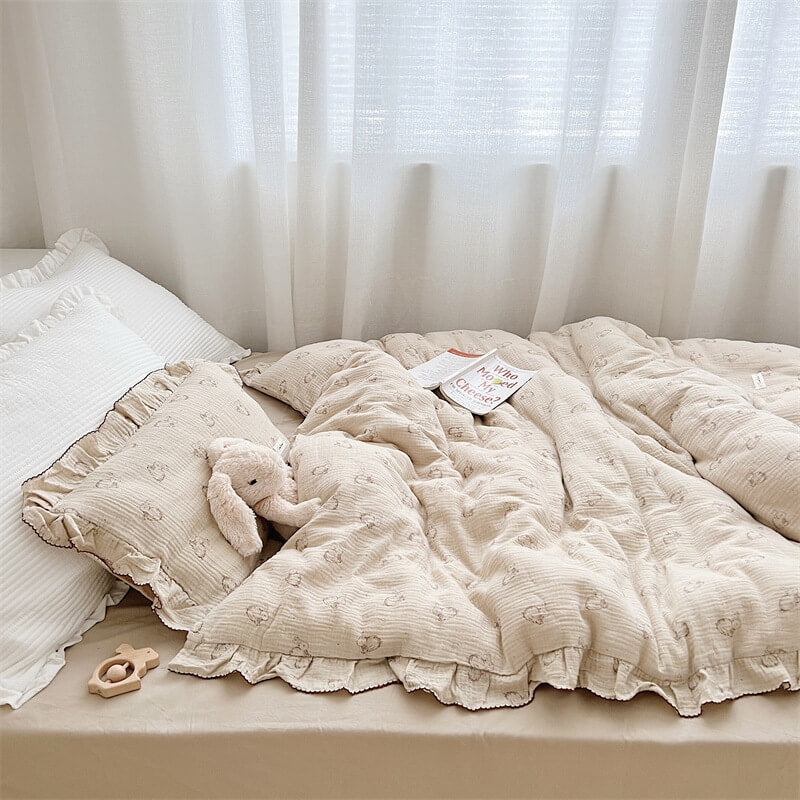 A side-view of a muslin duvet cover set, arranged on a twin-size bed. The bedding features a charming bunny print and ruffled edges with dark piping. The image also shows a white ruffled pillow, a plush teddy bear, and a book, highlighting the cozy and inviting feel of the bedding.