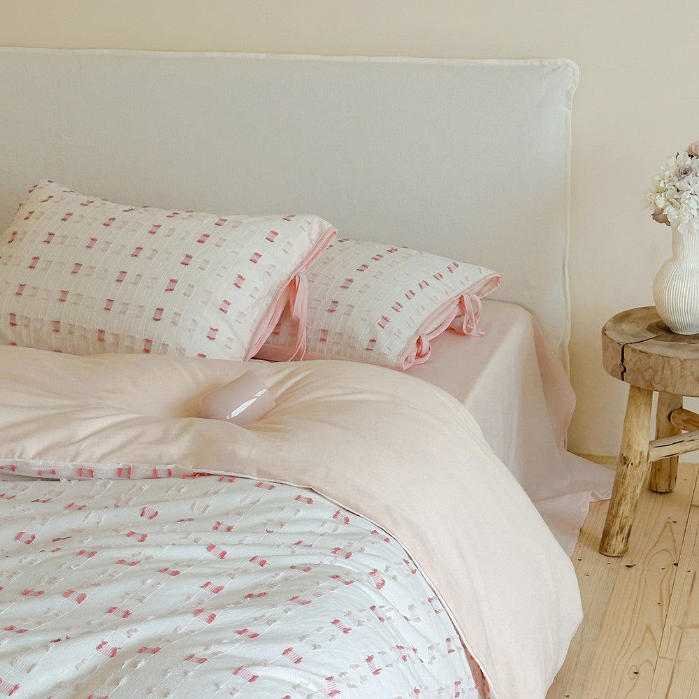Full shot of the Pastel Pink Tufted Check Cotton Bedding Set on a bed with a white linen headboard, showing the pink reverse of the duvet and the shams in a light-filled bedroom, next to a rustic wooden side table.
