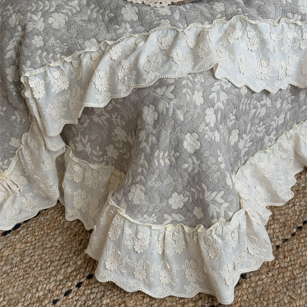 Detailed close-up of a grey quilted linen cotton bedspread showing the delicate white floral embroidery and the contrasting layered lace ruffle trim at the bottom edge, resting on a natural fiber rug.