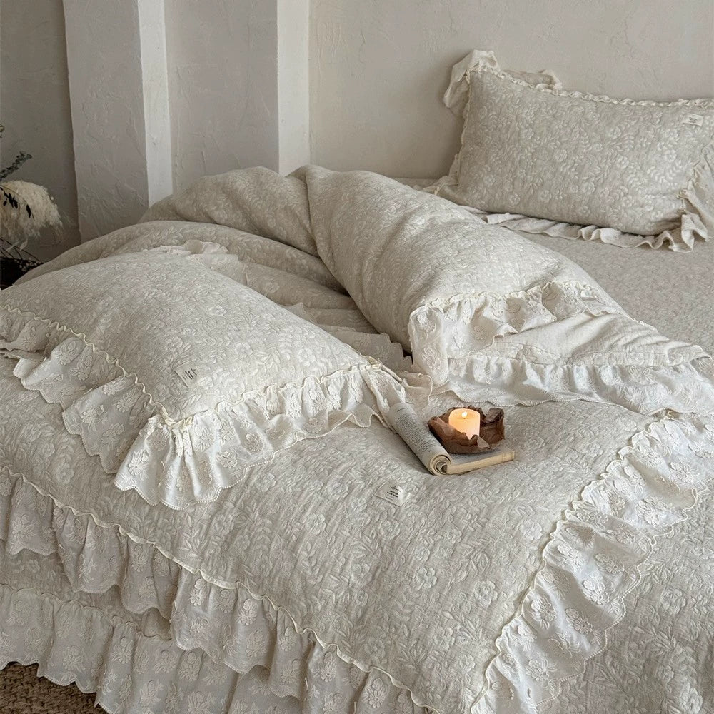 Close-up of a luxurious beige quilted linen cotton duvet cover and shams, featuring a subtle floral pattern and shabby chic ruffled French bedspread trim. The duvet is rumpled on a bed with a lit candle on a book, highlighting the cozy texture.