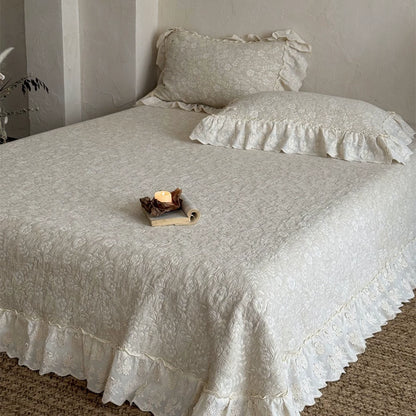 A wide shot of a neatly made bed covered with the beige quilted linen cotton bedspread. The French-style ruffle provides a floor-length dust ruffle effect, emphasizing the romantic, shabby chic aesthetic.