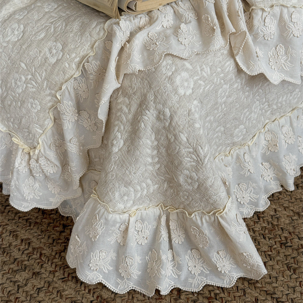 Extreme close-up of the beige quilted linen cotton bedspread detail, focusing on the textured floral quilt pattern and the multi-layered embroidered lace ruffle trim at the base of the bed.