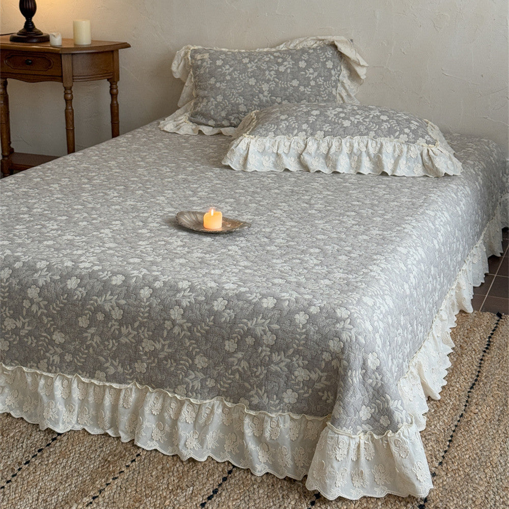 Full view of a twin bed styled with a grey quilted linen cotton bedspread with white floral pattern and shabby chic ruffled lace edge, paired with matching ruffled pillow shams. A lit candle sits on the bed.