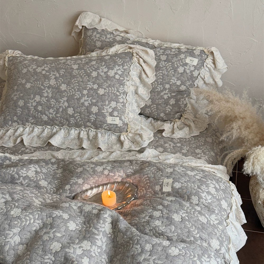 Close-up of the grey quilted linen cotton pillow shams and duvet cover, clearly showing the white floral pattern, the brand tags, and the decorative ruffle trim on the shams, styled with a candle.
